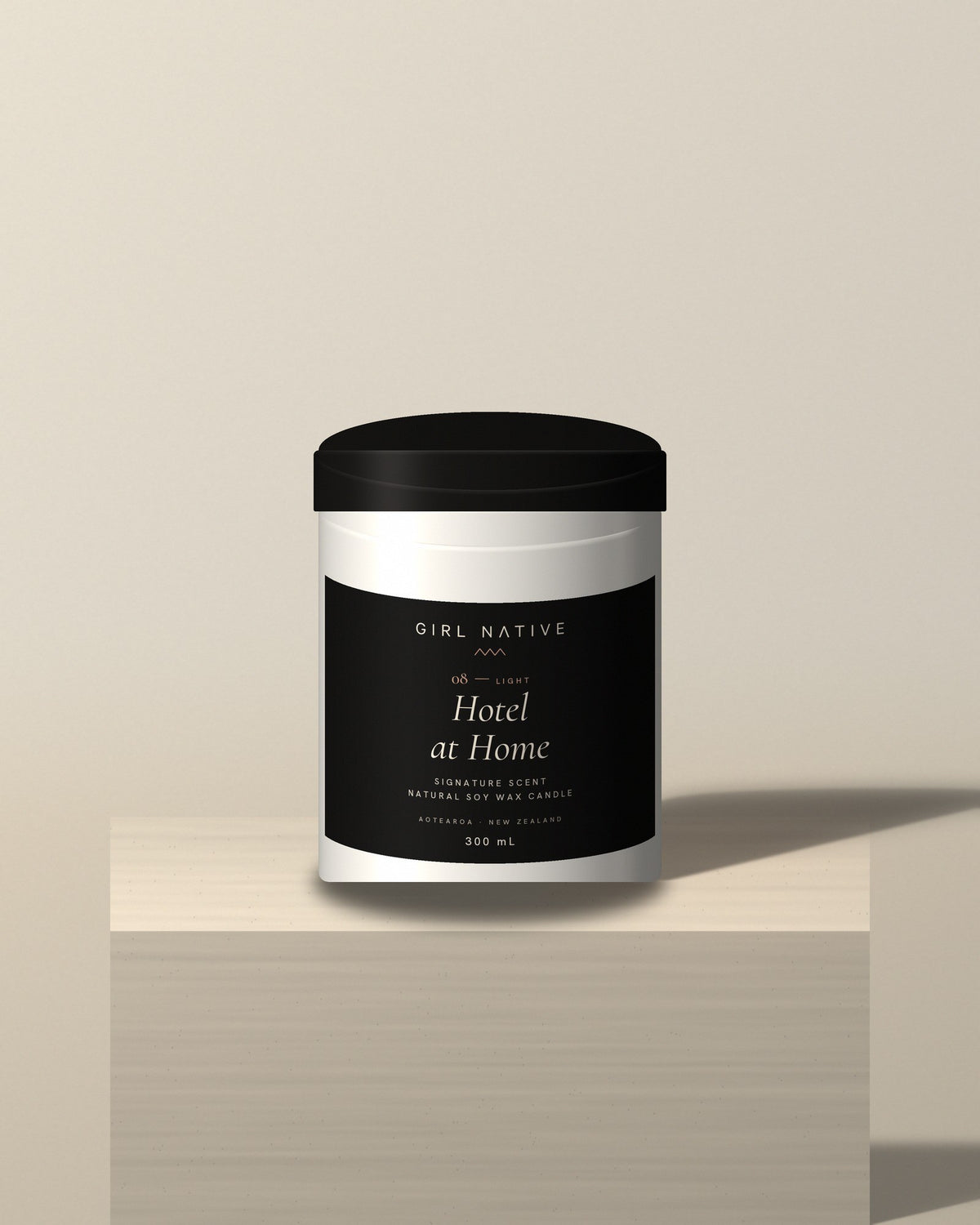 A cylindrical jar candle with a black lid and a black-and-white label reading 'GIRL NATIVE COFFEE Hotel at Home', displayed on a beige pedestal against a beige background.